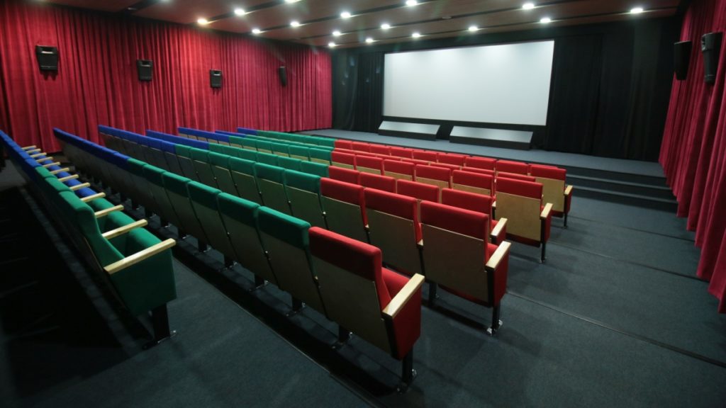 Puste kino z czerwonymi zasłonami, rzędami wyściełanych foteli w kolorach czerwonym, zielonym i niebieskim, podłogą pokrytą czarną wykładziną oraz dużym, pustym ekranem projekcyjnym z przodu. Świecą lampy sufitowe, widać też głośniki zamontowane na ścianach.