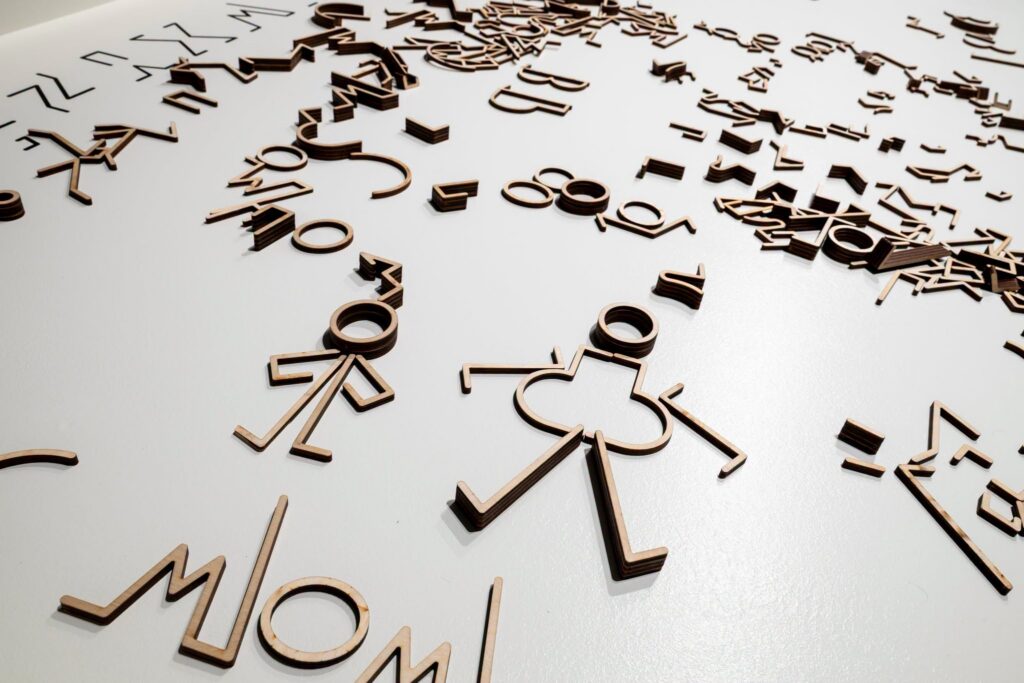 Wycinane laserowo drewniane elementy o różnych kształtach, w tym postacie w stylu „stick figure” i abstrakcyjne symbole, są rozrzucone po białej powierzchni – niektóre z nich tworzą skupiska, a inne są rozłożone.