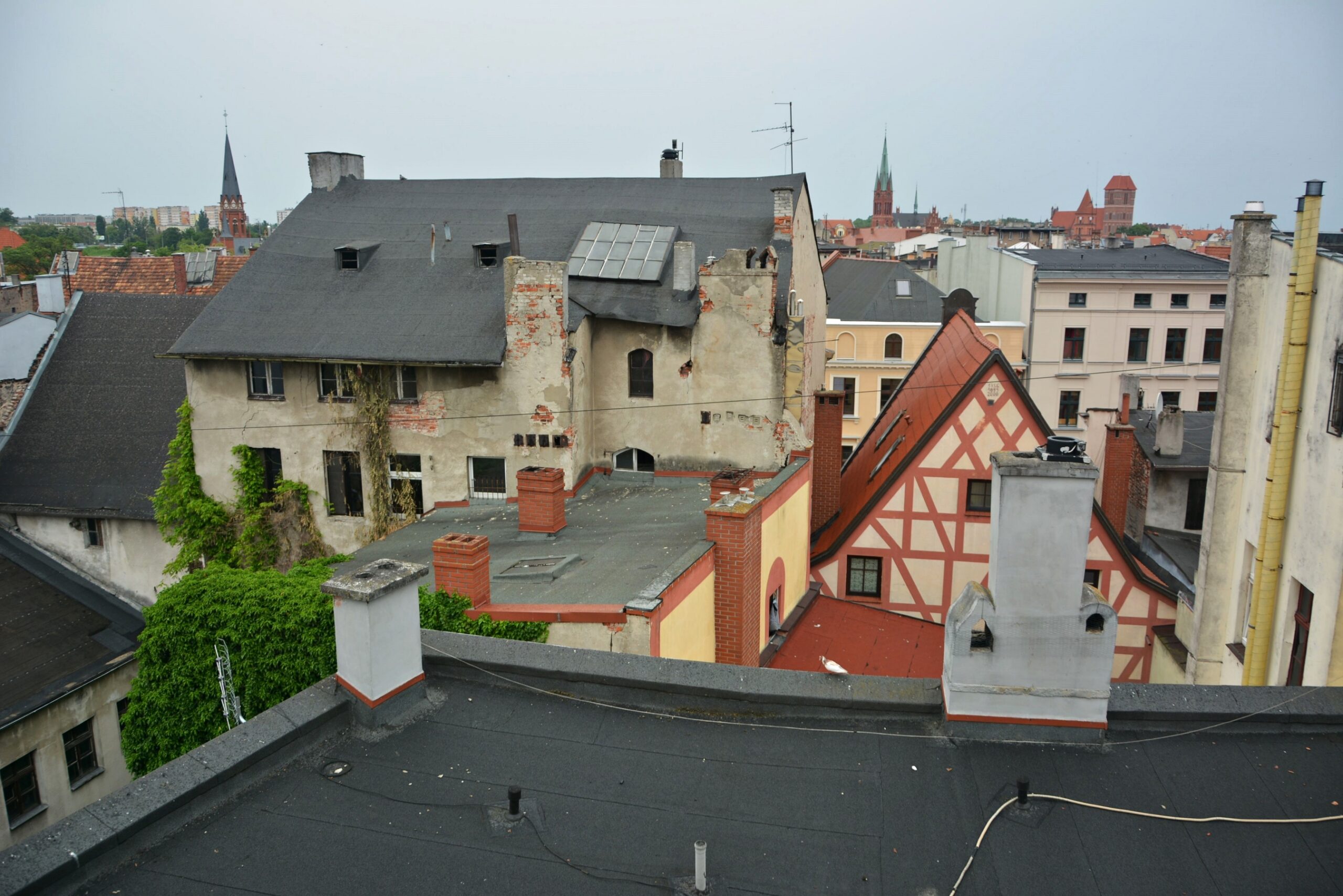 Widok z dachu przedstawiający mieszankę starych, zniszczonych przez czas budynków z widocznymi kominami i antenami. W tle, pod zachmurzonym niebem, widać wieże kościelne i kolejne dachy.