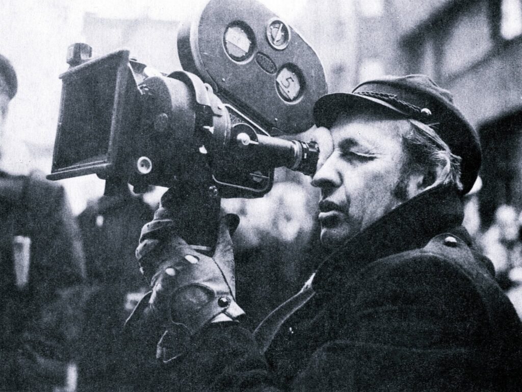 Osoba w czapce i rękawiczkach obsługuje na świeżym powietrzu zabytkowy aparat filmowy, przyłożywszy go do oka podczas kręcenia. Obraz jest czarno-biały.